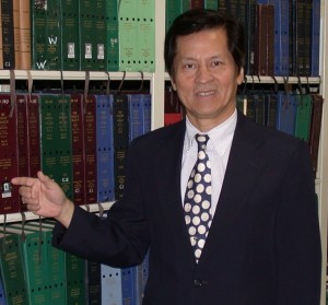 Image of man in dark suit, white shirt and necktie, standing in front of some bookshelves.