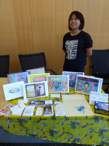 Dawn Wing wearing a black t-shirt standing in front of a table of zines.