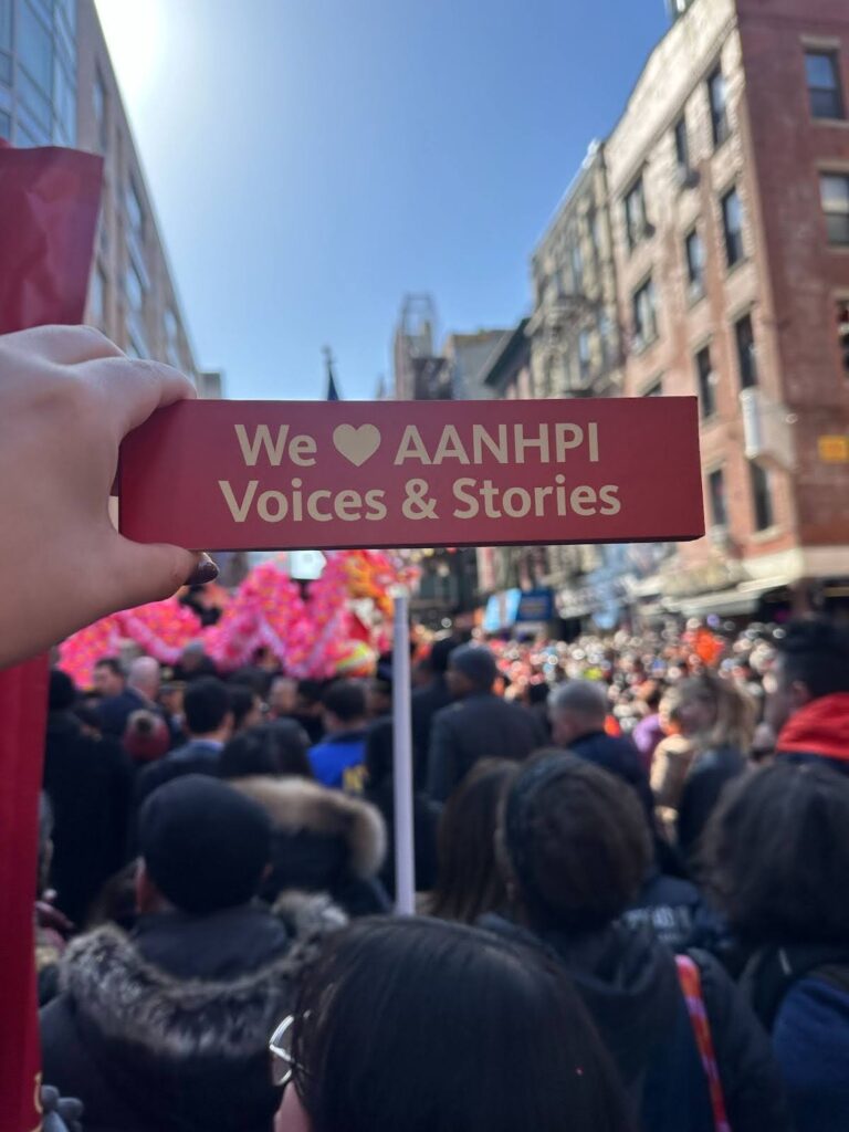 A hand holds a red bookmark above the parade crowd. The bookmark reads "We heart AANHPI voices and stories"