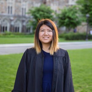 Christine Wei, female with straight shoulder length black-brown hair with blonde ends wearing an unzipped black graduation gown and royal blue top standing in a green space