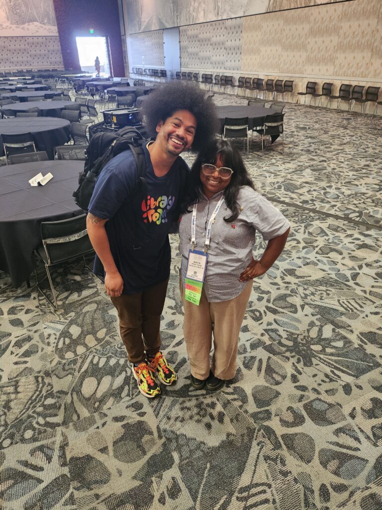 Joshlyn Thomas poses with library advocate and media personality, Mychael Threets. Mychael has a big afro and wears a navy shirt with the words "library joy" in rainbow colored font. Joshlyn has long black hair with bangs and white glasses