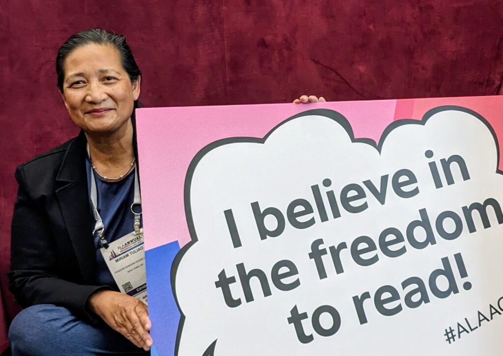 Mariam Tuliao smiles, wearing a black blazer and an ALA Annual Conference badge. She holds a sign that reads "I believe in the freed to read" in a comic book style speech bubble.