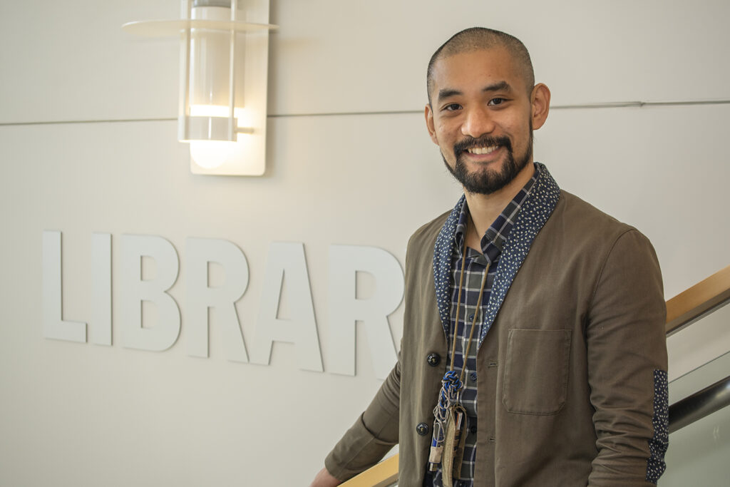Di Zhang smiles at the camera. He has close-shaved black hair and a clean black beard. He wears a brown suit jacket with polka dot lapels and elbow patches over a dark plaid button up. Behind him, the word "LIBRARY" is plastered on the wall beneath a wall lamp.