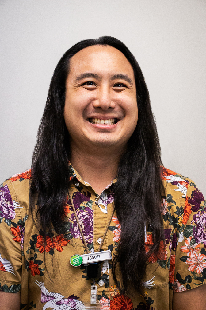 Jason smiles brightly at a point just above the viewer. He has long dark hair. He wears a floral button up that is yellow with accents of orange, purple, and white.