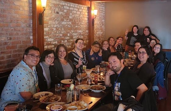 Fifteen members of the APALA Pacific Northwest Chapter smile around a table laden with food. The wall behind them is exposed brick with two hanging lanterns for cozy ambience.