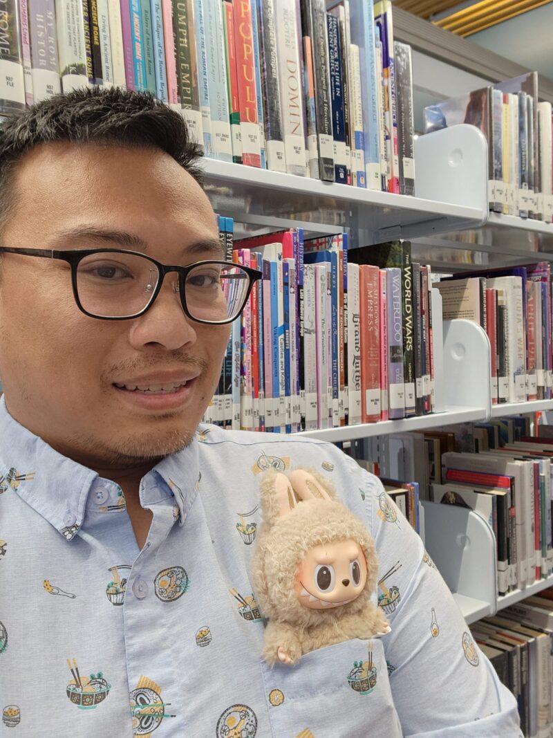 Librarian Warren Chin, wearing glasses and a light blue shirt, with a tan Labubu in his left shirt pocket.